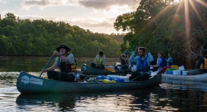 A group of students paddle canoes on a river surrounded by green trees.